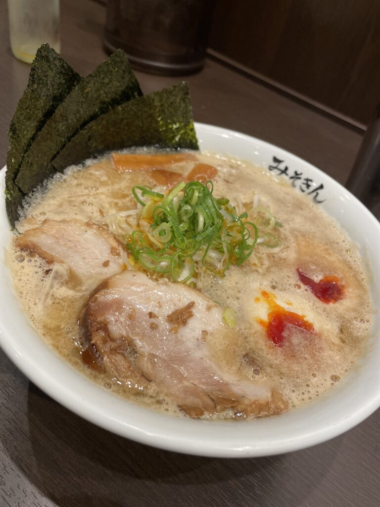 A bowl of ramen featuring slices of pork, green onions, and toppings, accompanied by sheets of nori.