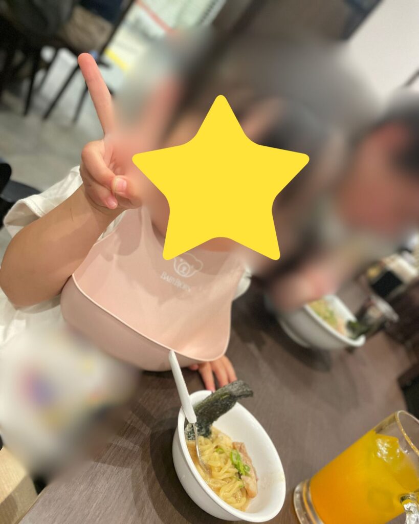 子どもがラーメンを食べながらピースサインをしている写真。テーブルにはラーメンと飲み物が置かれています。