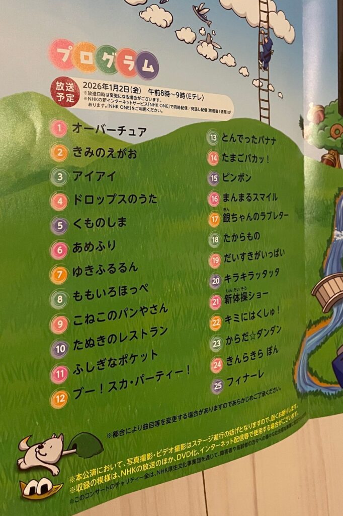 ファミリーコンサートのプログラムリストが描かれた紙。タイトルは「プログラム」で、収録される曲のリストがカラフルなフォントで表示されている。背景には緑の草原と青空、白い雲が描かれている。